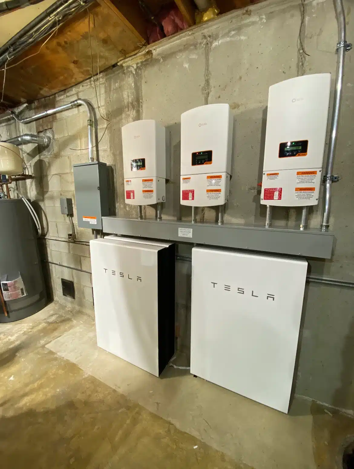 Tesla Powerwall battery storage units installed below three solar inverters on a concrete basement wall for home energy backup and management.